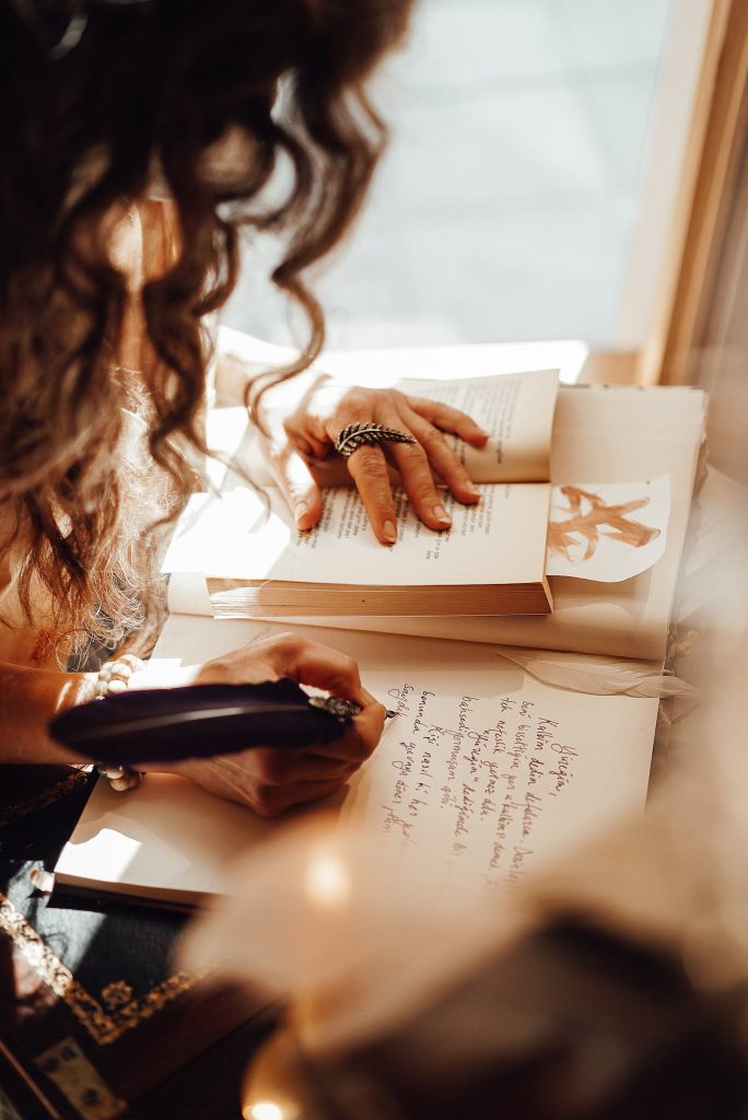 Crop unrecognizable female with curly hair taking notes with ink while sitting at vintage writing cabinet in retro style store with writing supplies
