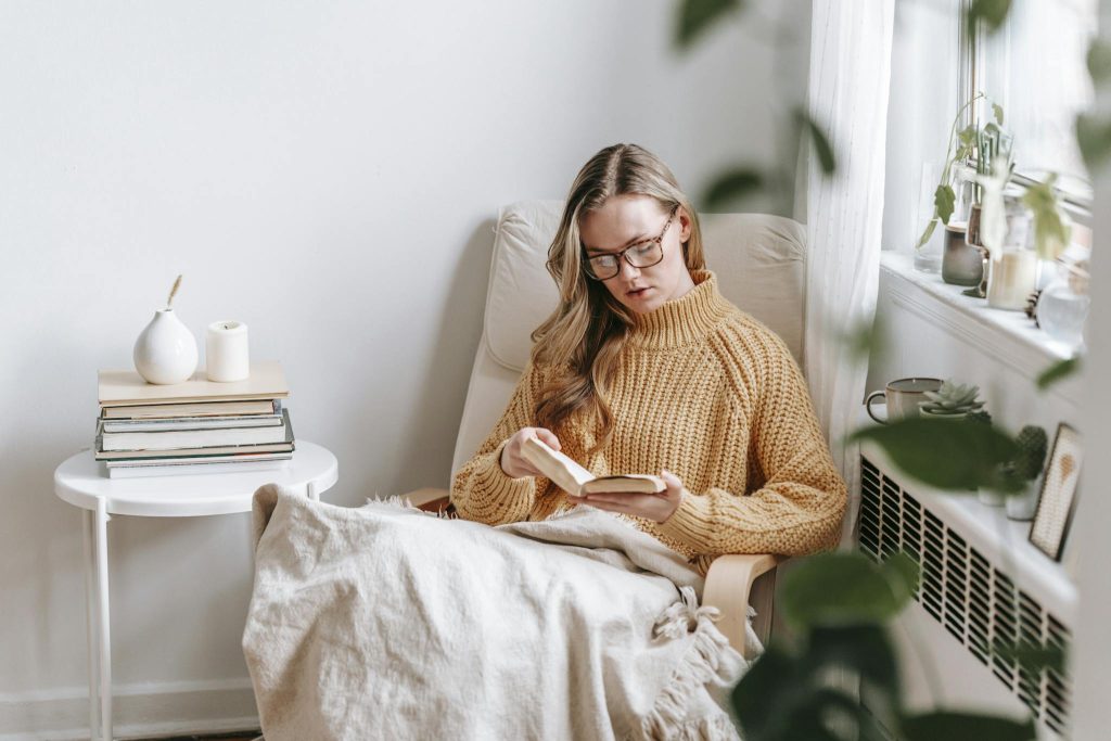 Young female student in trendy sweater and glasses sitting in white armchair with plaid near table with books and candle while reading book